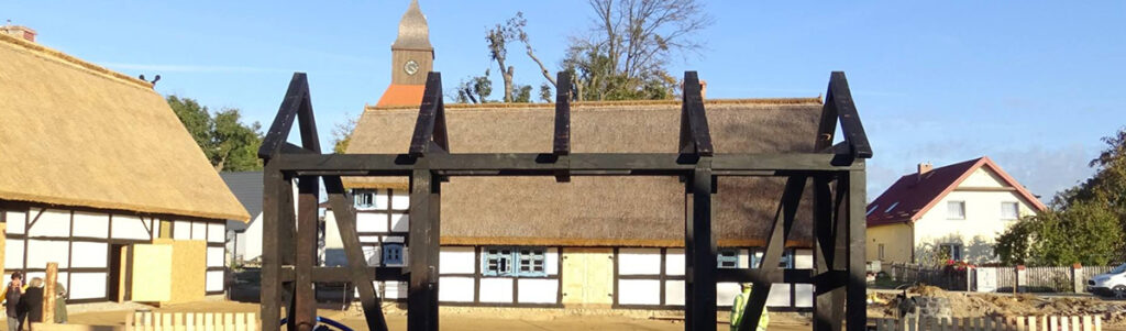 Zagroda Jamneńska: drewniana konstrukcja na pierwszym planie, w tle rekonstrukcje historycznych domów szachulcowych z dachami ze strzechy oraz wieża kościoła. Słoneczny dzień.