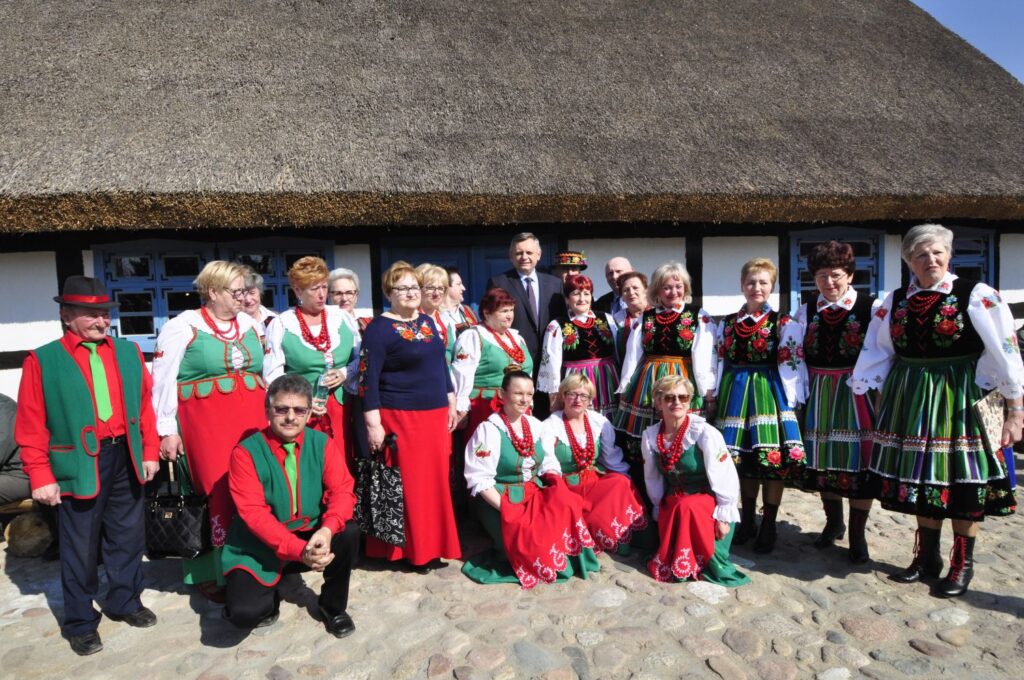 Liczna grupa osób w kolorowych, tradycyjnych strojach ludowych (jamneńskich) pozuje do zdjęcia przed zabytkową chatą krytą strzechą (kliknięcie powiększy zdjęcie).