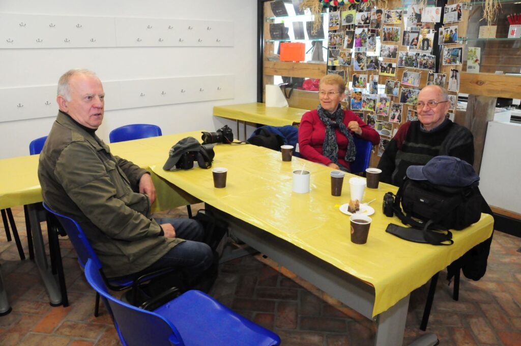 Grupa dorosłych fotografów siedzi przy stole w sali warsztatowej, rozmawiając przy kawie podczas spotkania (kliknięcie powiększy zdjęcie).