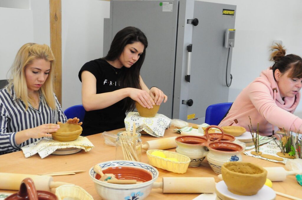 Kilka osób skupionych na lepieniu naczyń z gliny przy długim stole warsztatowym, na którym leżą wałki, miski i przybory ceramiczne (kliknięcie powiększy zdjęcie).