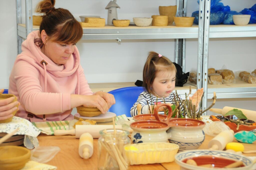 Kobieta i małe dziecko wspólnie uczestniczą w warsztatach ceramicznych, lepiąc gliniane naczynie przy stole pełnym narzędzi (kliknięcie powiększy zdjęcie).
