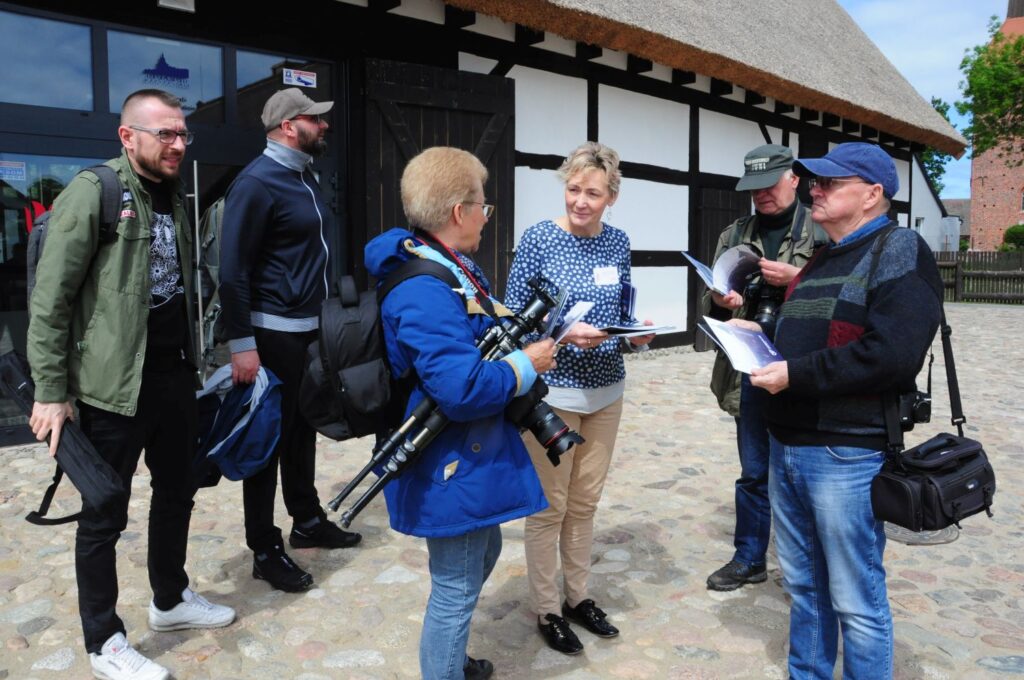 Fotografowie stoją na dziedzińcu przy zabytkowym budynku w trakcie rozmowy i przygotowań do wspólnego działania, część osób trzyma sprzęt fotograficzny (kliknięcie powiększy zdjęcie).