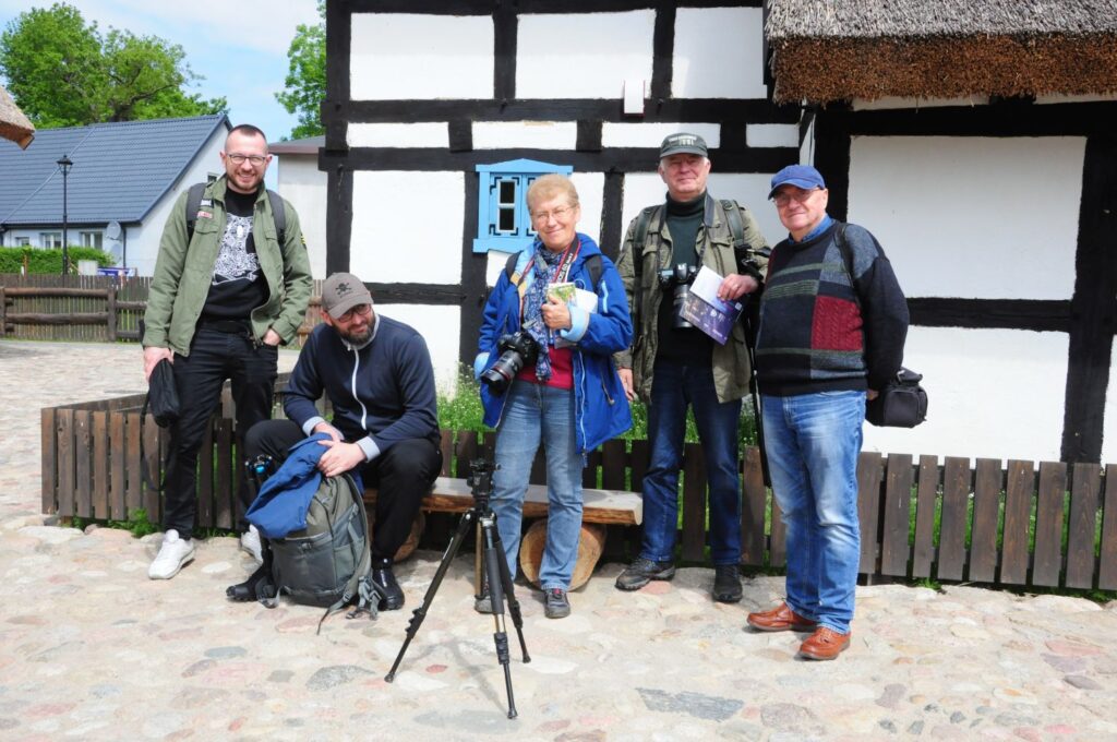 Osoby biorące udział w plenerze fotograficznym pozują do zdjęcia przed budynkiem Zagrody, obok statywu i aparatów fotograficznych, w trakcie wydarzenia plenerowego (kliknięcie powiększy zdjęcie).