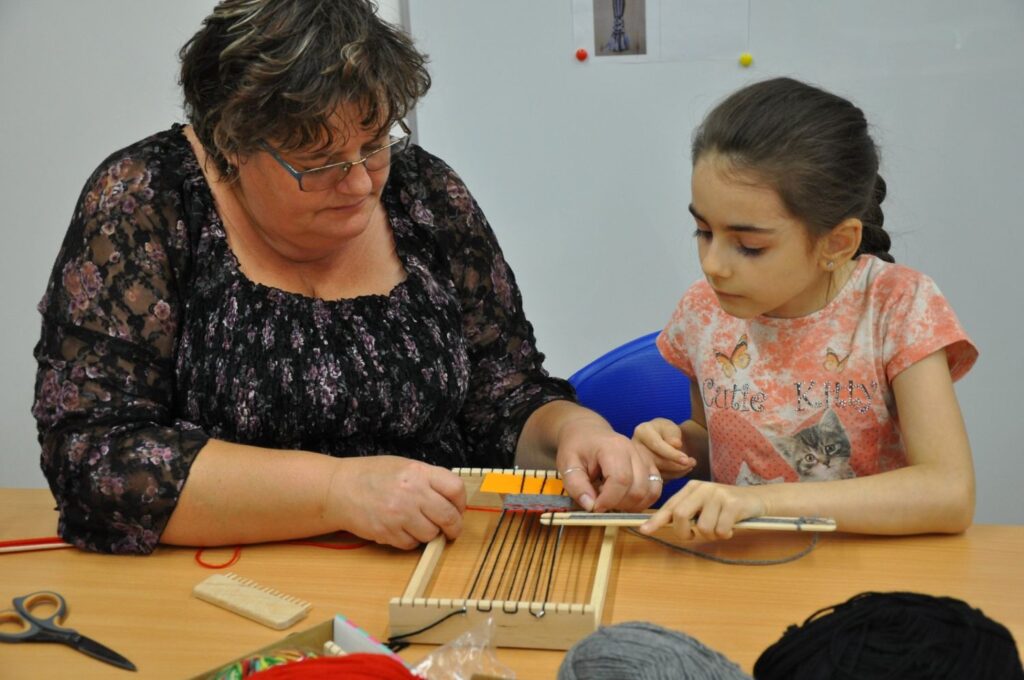 Kobieta pomaga dziewczynce podczas tkania kolorowych włóczek na małych krosnach stołowych w trakcie warsztatów tkackich (kliknięcie powiększy zdjęcie).