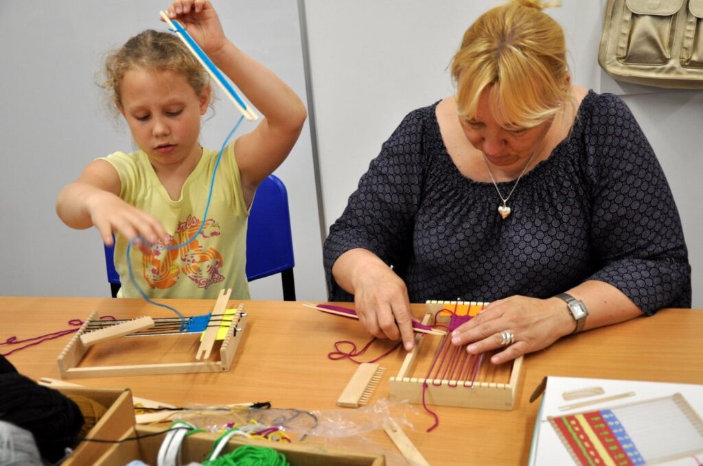 Dziecko i dorosła uczestniczka warsztatów tkackich pracują przy krosnach, przeplatając barwne nici włóczki (kliknięcie powiększy zdjęcie).