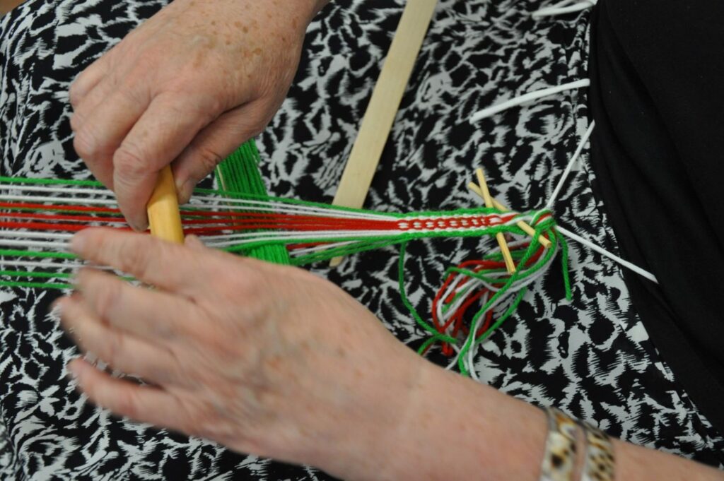 Zbliżenie na dłonie uczestniczki warsztatów tkania na bardkach, układającej zielone i czerwone nici wzoru (kliknięcie powiększy zdjęcie).