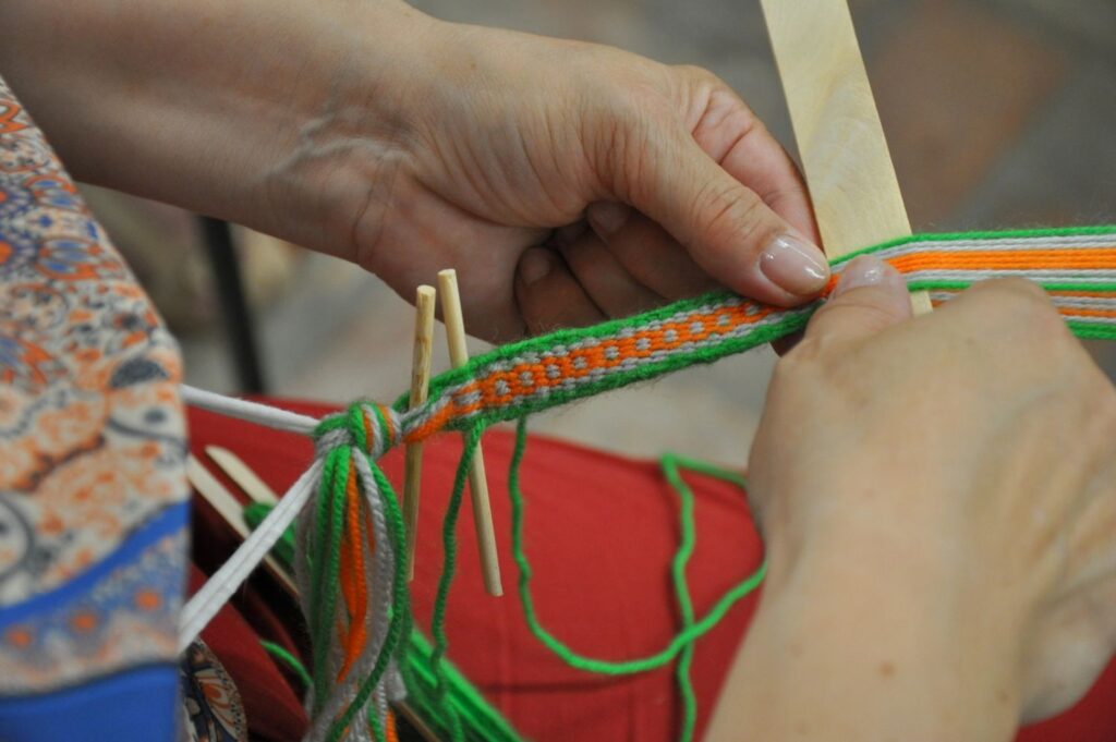 Zbliżenie na dłonie uczestniczki warsztatów tkania na bardkach, układającej kolorowe nici wzoru (kliknięcie powiększy zdjęcie).