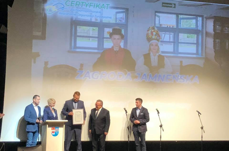 Pięć osób stojących na scenie. Po środku osoba pełniąca obowiązki dyrektora Muzeum w Koszalinie, odbiera certyfikat, w tle na ekranie widoczny napis „Zagroda Jamneńska” oraz zdjęcie osób w strojach ludowych.