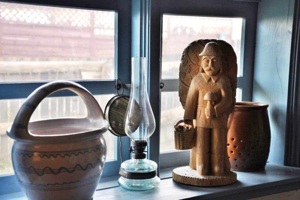 Drewniana rzeźba mężczyzny w kapeluszu trzymającego koszyk z grzybami, ustawiona na parapecie obok glinianego naczynia i lampy naftowej, w tle okno z widokiem na zabudowania (kliknięcie powiększy zdjęcie).