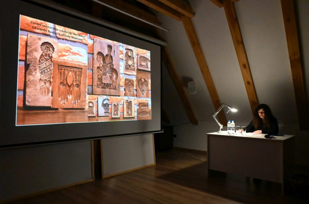 Prelegentka siedzi przy biurku obok dużego ekranu z prezentacją przedstawiającą zabytkowe przedmioty związane z tematyką kulinarną (kliknięcie powiększy zdjęcie).