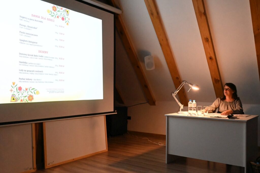 Prelegentka siedzi przy biurku obok ekranu z przykładowym menu dań dla dzieci oraz deserów i prezentuje kolejne zagadnienia seminarium (kliknięcie powiększy zdjęcie).