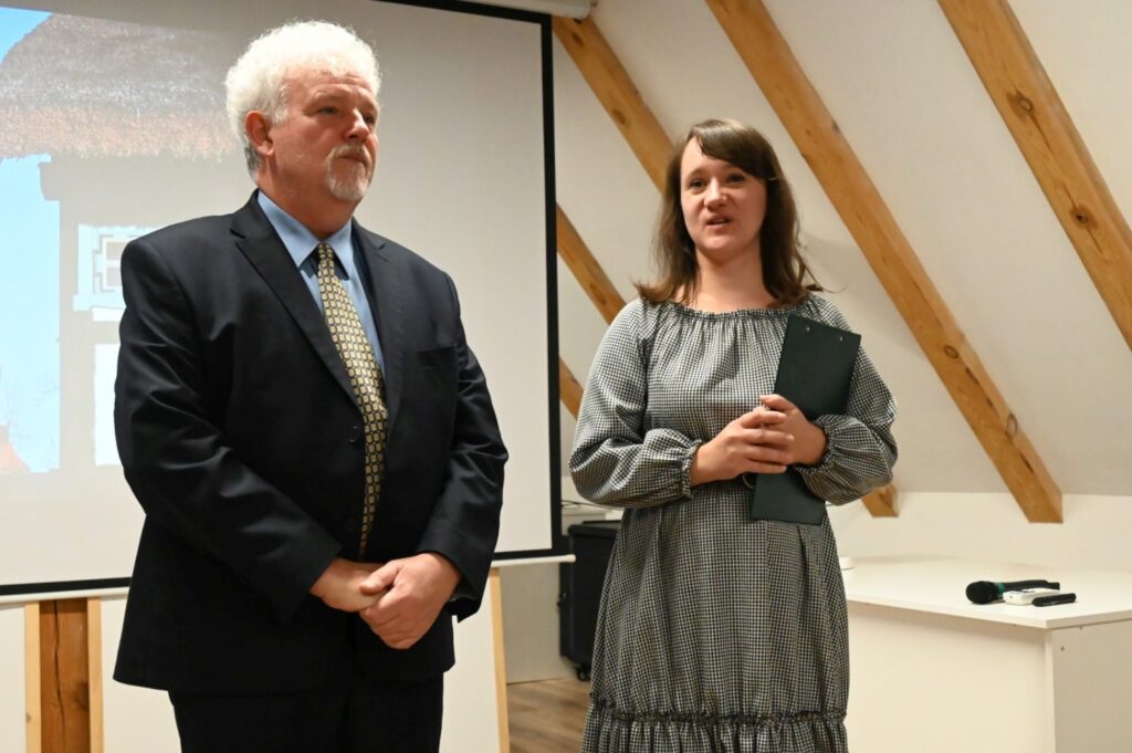 Kobieta i mężczyzna stoją przed ekranem projekcyjnym w sali z drewnianymi belkami stropowymi, otwierając seminarium (kliknięcie powiększy zdjęcie).