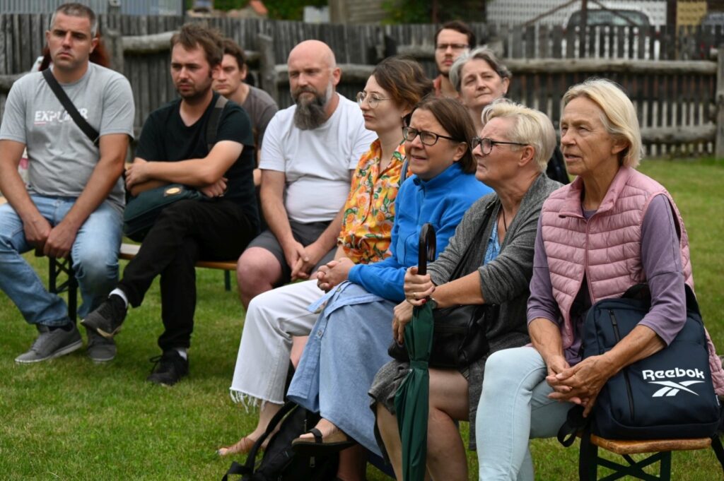 Zbliżenie na grupę słuchaczy siedzących na ławkach podczas wykładu w plenerze, skupionych na wypowiedzi prowadzącego (kliknięcie powiększy zdjęcie).