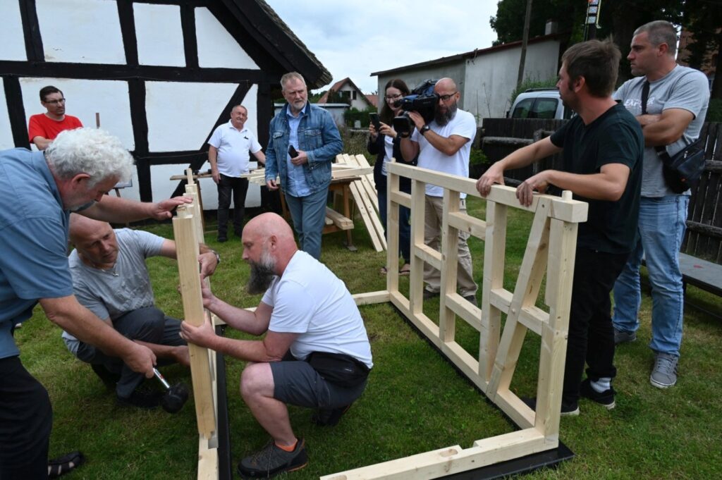 Kilku mężczyzn montuje pionowy element drewnianej konstrukcji, przytrzymując belki i dopasowując je do podstawy, w tle obserwatorzy i kamera (kliknięcie powiększy zdjęcie).