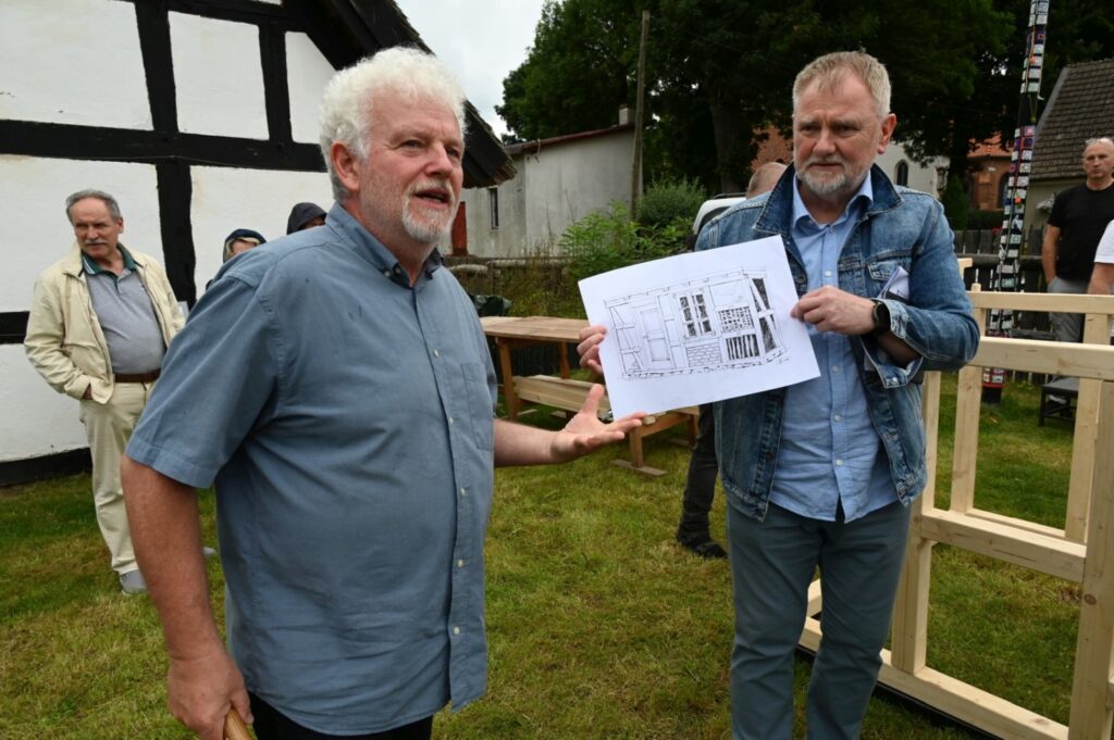 Dwóch mężczyzn stoi na trawie obok drewnianej konstrukcji, jeden z nich prezentuje planszę z rysunkiem projektu podczas spotkania na świeżym powietrzu (kliknięcie powiększy zdjęcie).