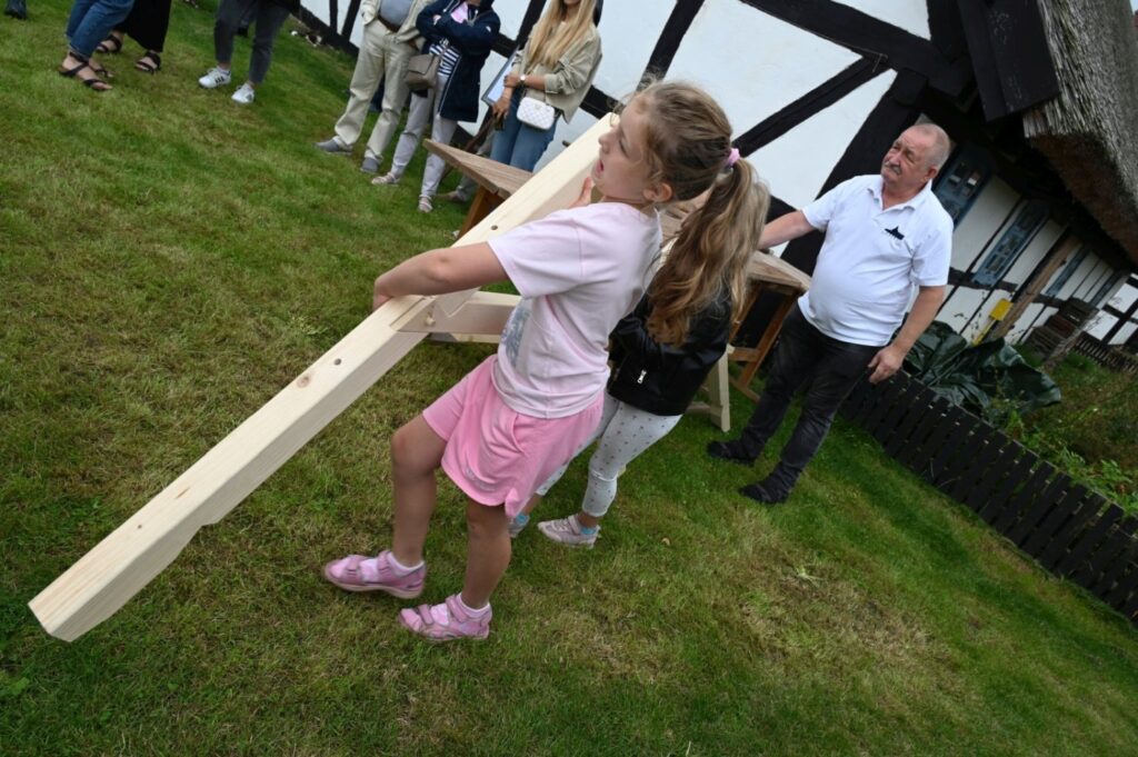 Dziewczynka niesie długą drewnianą belkę po trawie w kierunku powstającej konstrukcji, obok stoją dorośli uczestnicy warsztatów (kliknięcie powiększy zdjęcie).