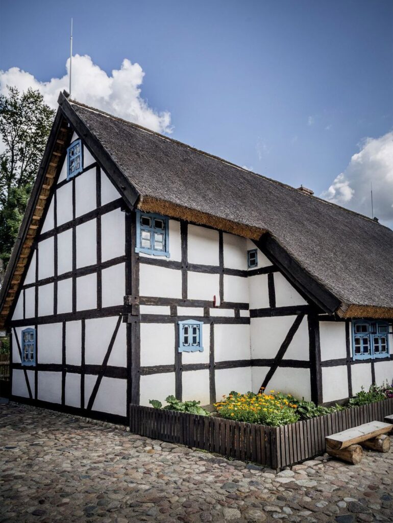 Rekonstrukcja zabytkowego domu szachulcowego z dachem krytym strzechą, biało-czarną elewacją i niebieskimi okiennicami, stojący przy brukowanym dziedzińcu z rabatą kwiatową (kliknięcie powiększy zdjęcie).