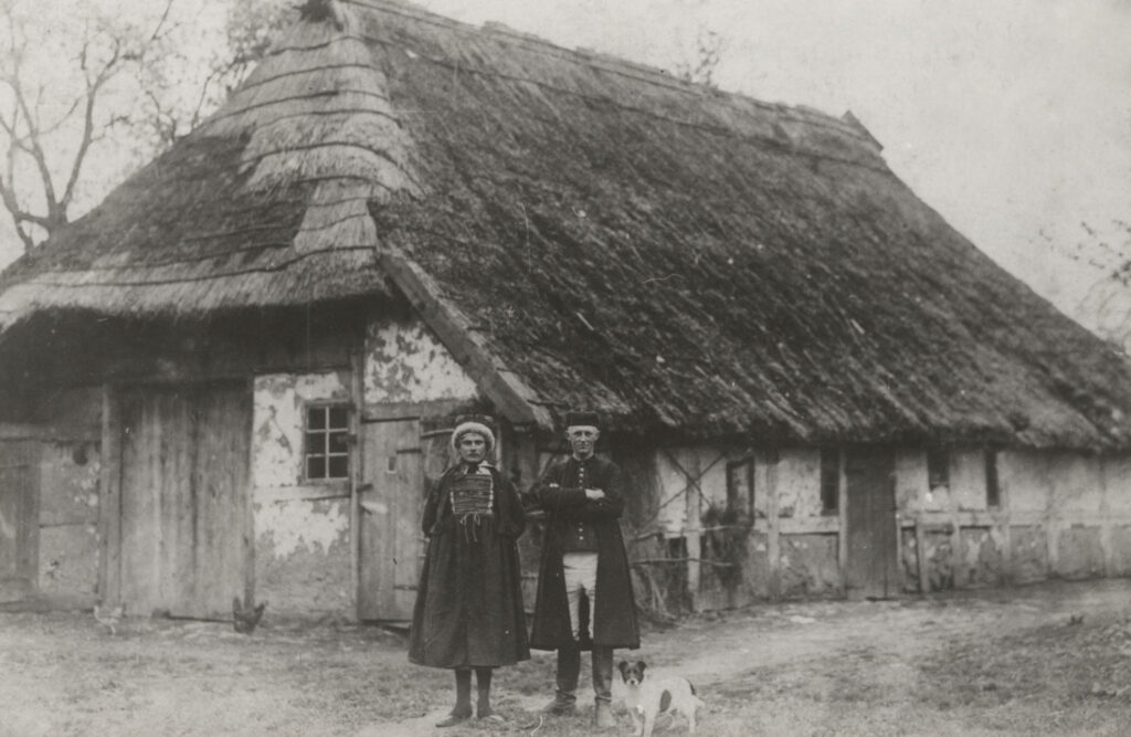 Czarno-białe archiwalne zdjęcie przedstawiające kobietę i mężczyznę stojących przed wiejską chałupą krytą strzechą, ubranych w tradycyjne stroje, u ich stóp siedzi mały pies, a w tle widoczna jest ściana budynku z małym oknem i drewnianymi drzwiami (kliknięcie powiększy zdjęcie).