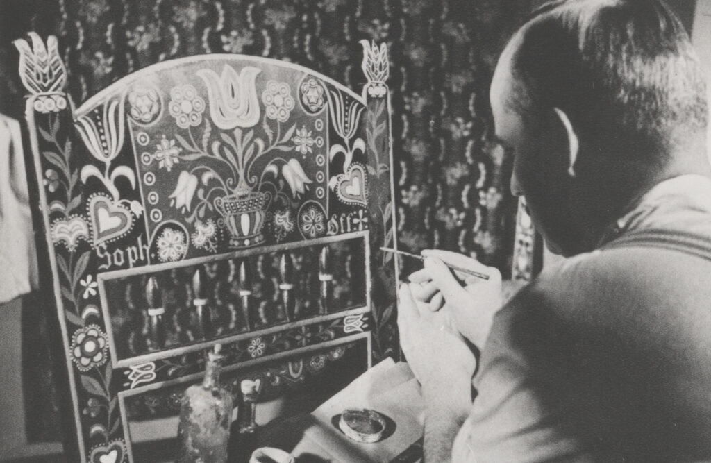 Czarno-białe zdjęcie ukazujące mężczyznę malującego bogato zdobione oparcie krzesła z jamneńskimi motywami kwiatowymi i sercami, siedzącego przy stole z farbami, na tle dekoracyjnej tkaniny (kliknięcie powiększy zdjęcie).