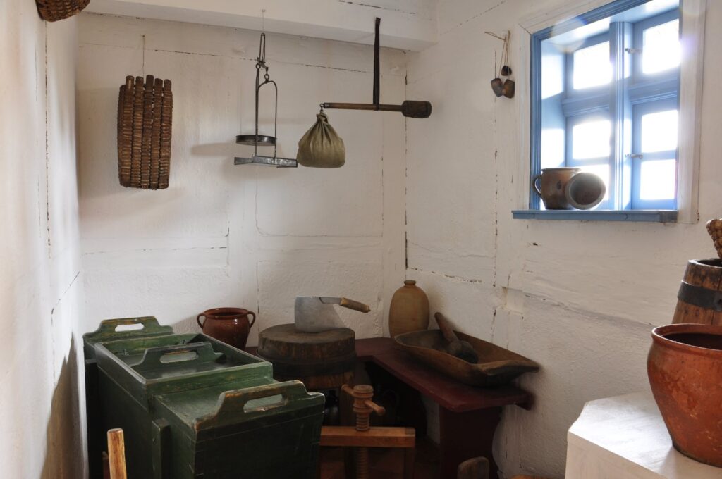 Wnętrze dawnej kuchni z naczyniami, wagą i sprzętami gospodarczymi umieszczonymi przy ścianach (kliknięcie powiększy zdjęcie).