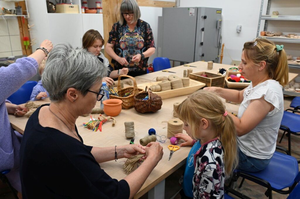 Warsztaty rękodzielnicze, podczas których dorośli i dzieci wspólnie wykonują prace z gliny przy stole warsztatowym (kliknięcie powiększy zdjęcie).