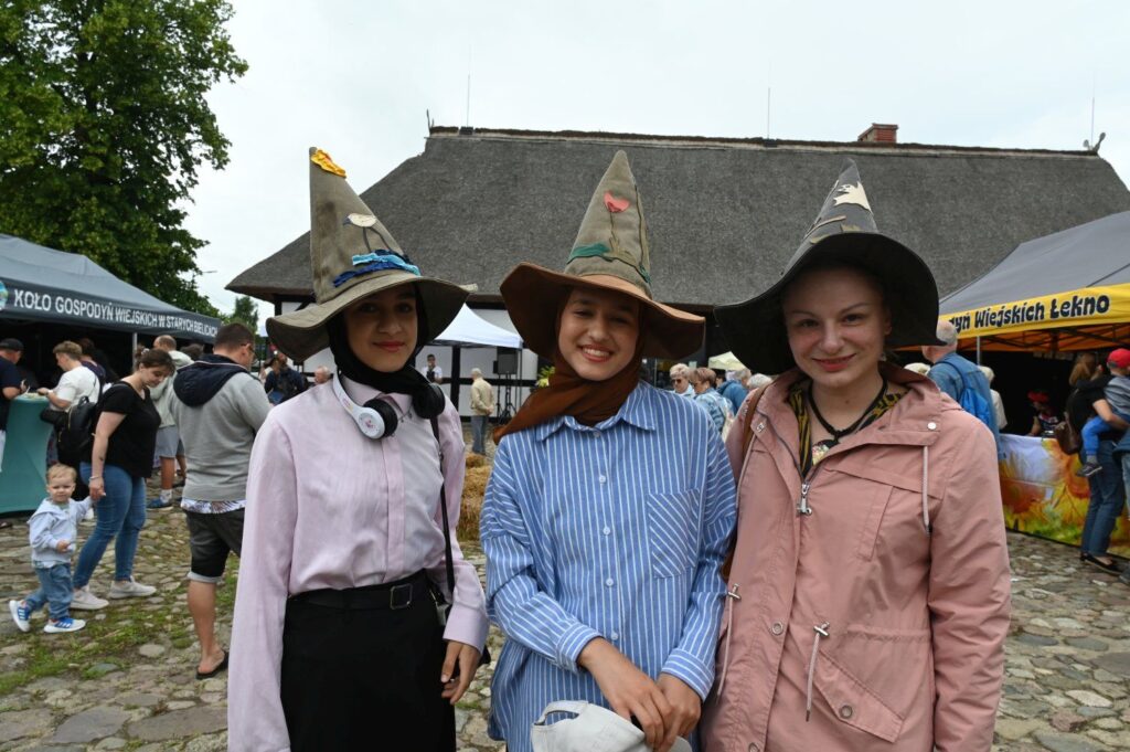 Trzy dziewczęta w kapeluszach pozujące do zdjęcia na tle stoisk jarmarku jamneńskiego (kliknięcie powiększy zdjęcie).