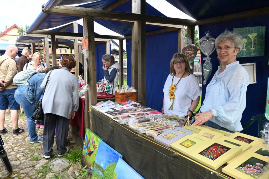 Wystawczynie stojące za stoiskiem z rękodziełem i grafikami, rozmawiające z odwiedzającymi (kliknięcie powiększy zdjęcie).