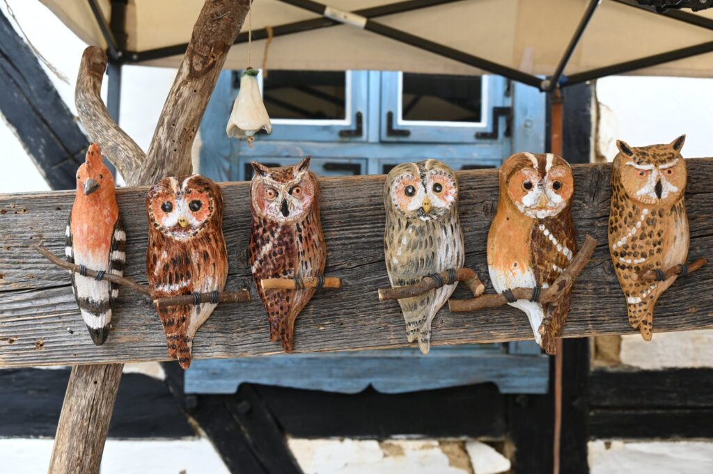 Ręcznie wykonane ceramiczne figurki sów ustawione na drewnianej belce na stoisku artystycznym (kliknięcie powiększy zdjęcie).