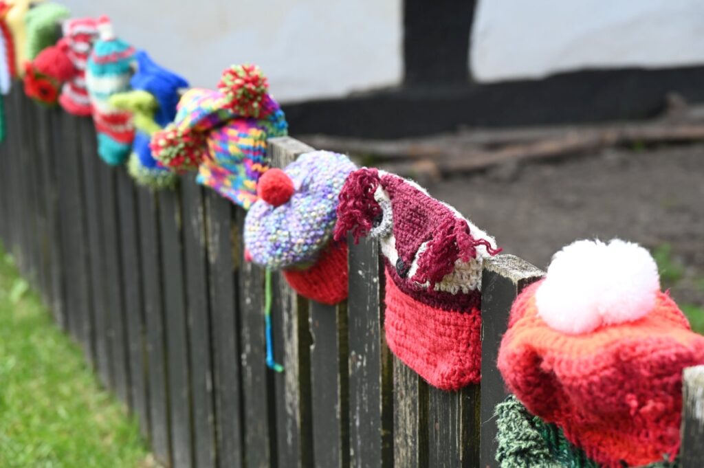 Kolorowe, dziergane czapki dziecięce zawieszone na drewnianym płocie jako ekspozycja plenerowa (kliknięcie powiększy zdjęcie).