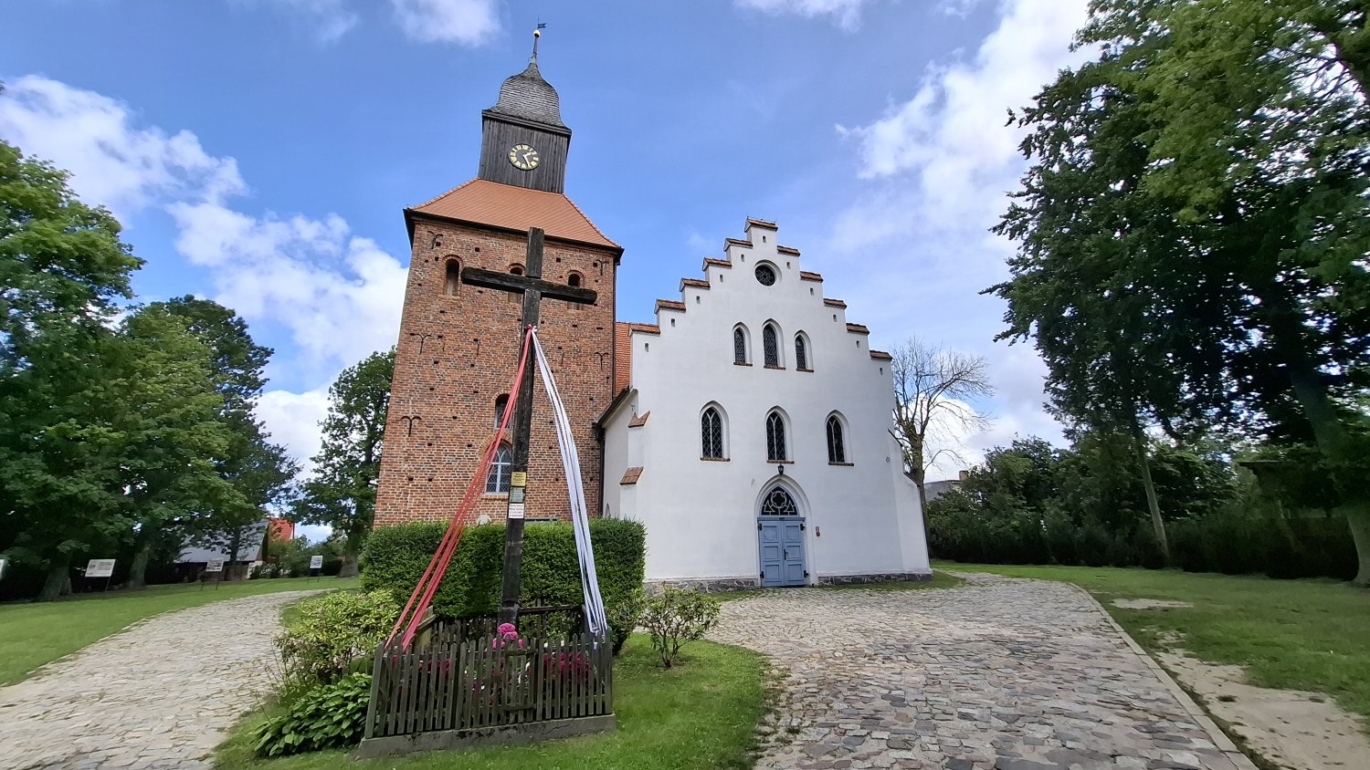 Widok na kościół w Jamnie od strony południowej – biała fasada, ceglana wieża oraz niewielka drewniany krzyż przy wejściu, otoczenie zieleni i brukowana nawierzchnia (kliknięcie powiększy zdjęcie).