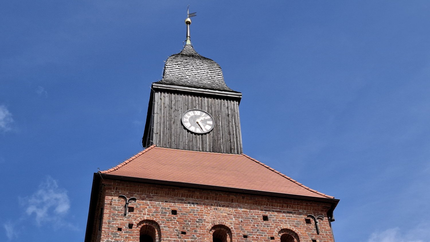 Zbliżenie na wieżę kościoła w Jamnie – drewniana konstrukcja z zegarem oraz hełm wieży zwieńczony metalowym krzyżem, na tle czystego, niebieskiego nieba (kliknięcie powiększy zdjęcie).