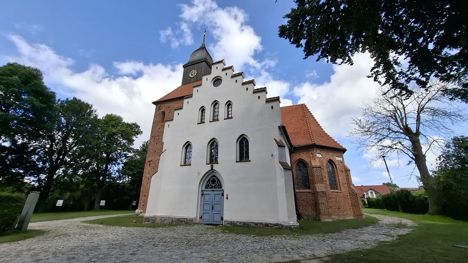 Frontowy widok kościoła w Jamnie z białą, schodkową elewacją szczytową i ceglaną wieżą, przed wejściem brukowana ścieżka, wokół drzewa i trawa (kliknięcie powiększy zdjęcie).