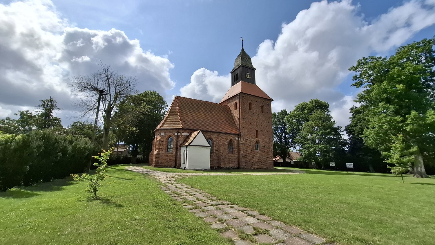 Widok na zabytkowy kościół w Jamnie z boku, ceglany korpus świątyni z wieżą, prowadząca do niego kamienna ścieżka oraz zielony trawnik, w tle drzewa i częściowo zachmurzone niebo (kliknięcie powiększy zdjęcie).