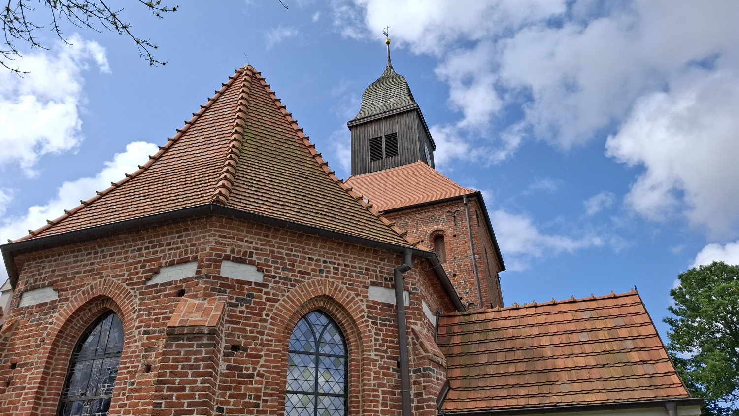 Detal architektoniczny kościoła w Jamnie – ceglana ściana prezbiterium z ostrołukowym oknem oraz fragment dachu, w tle wieża kościelna i błękitne niebo z chmurami (kliknięcie powiększy zdjęcie).