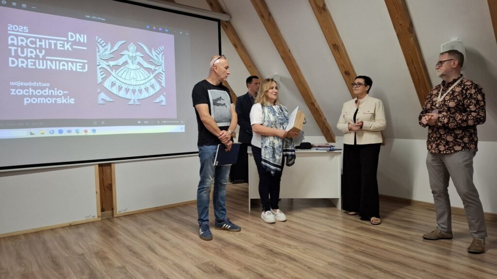 Prelegenci stojący przed ekranem projekcyjnym podczas otwarcia wydarzenia „Dni Architektury Drewnianej”, widoczna prezentacja na ścianie (kliknięcie powiększy zdjęcie)