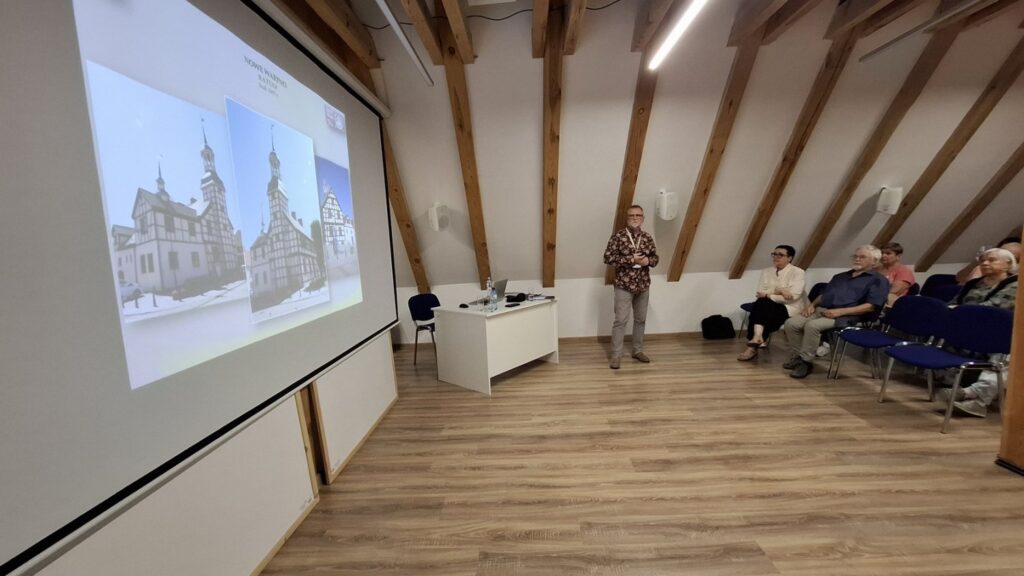 Slajd z fotografiami zabytkowych budynków wyświetlany na ekranie podczas wykładu, prelegent i słuchacze w sali (kliknięcie powiększy zdjęcie)
