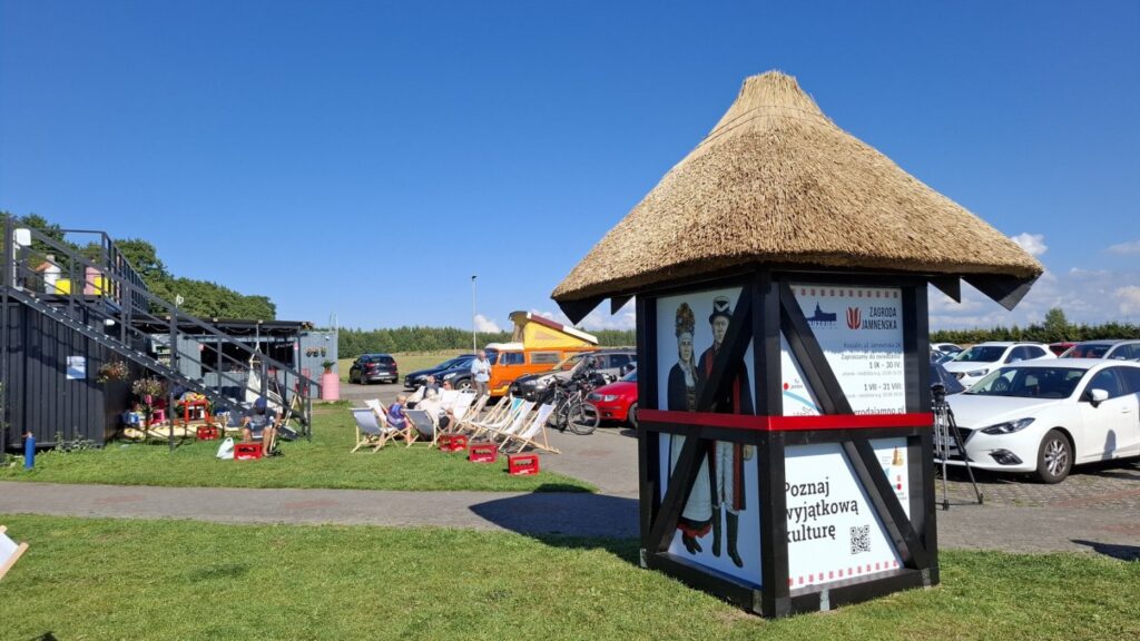 Witacz jamneński stoi na terenie zielonym, w tle zaparkowane samochody a na trawniku leżaki (kliknięcie powiększy zdjęcie)