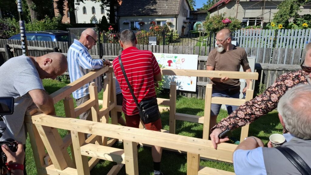 Uczestnicy wspólnie skręcają drewniane elementy konstrukcji na terenie zielonym, w tle zabudowania Jamna (kliknięcie powiększy zdjęcie)