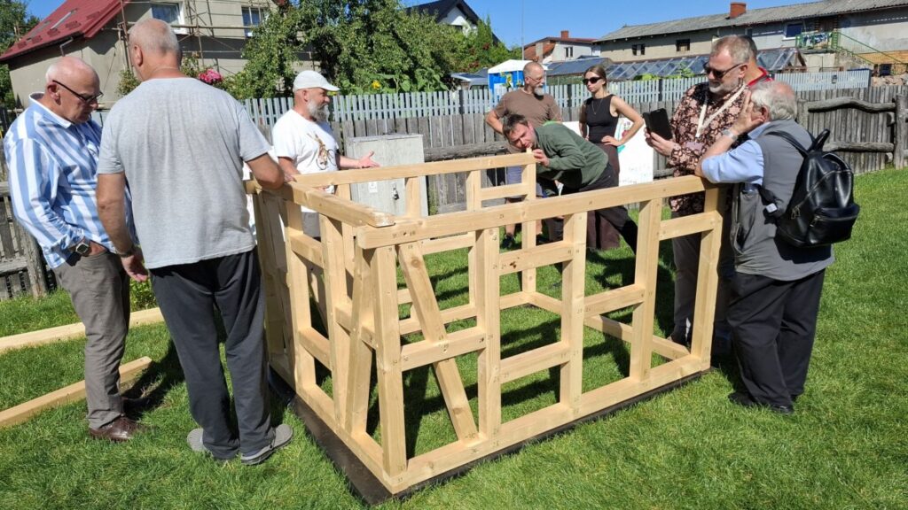 Uczestnicy wspólnie skręcają drewniane elementy konstrukcji na terenie zielonym, w tle zabudowania Jamna (kliknięcie powiększy zdjęcie)