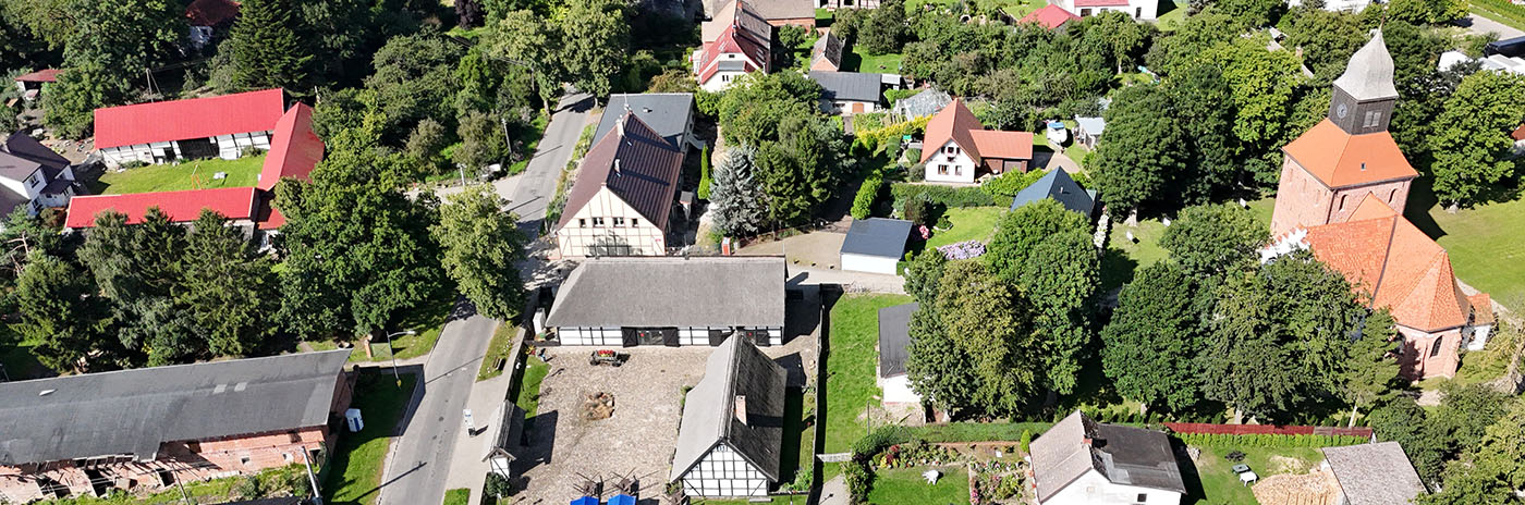 Widok z lotu ptaka na Jmano, w centrum znajdują się zabytkowe domy z muru pruskiego, a po prawej stronie ceglany kościół z wysoką wieżą. Całość otoczona jest gęstą zielenią drzew i ogrodów.