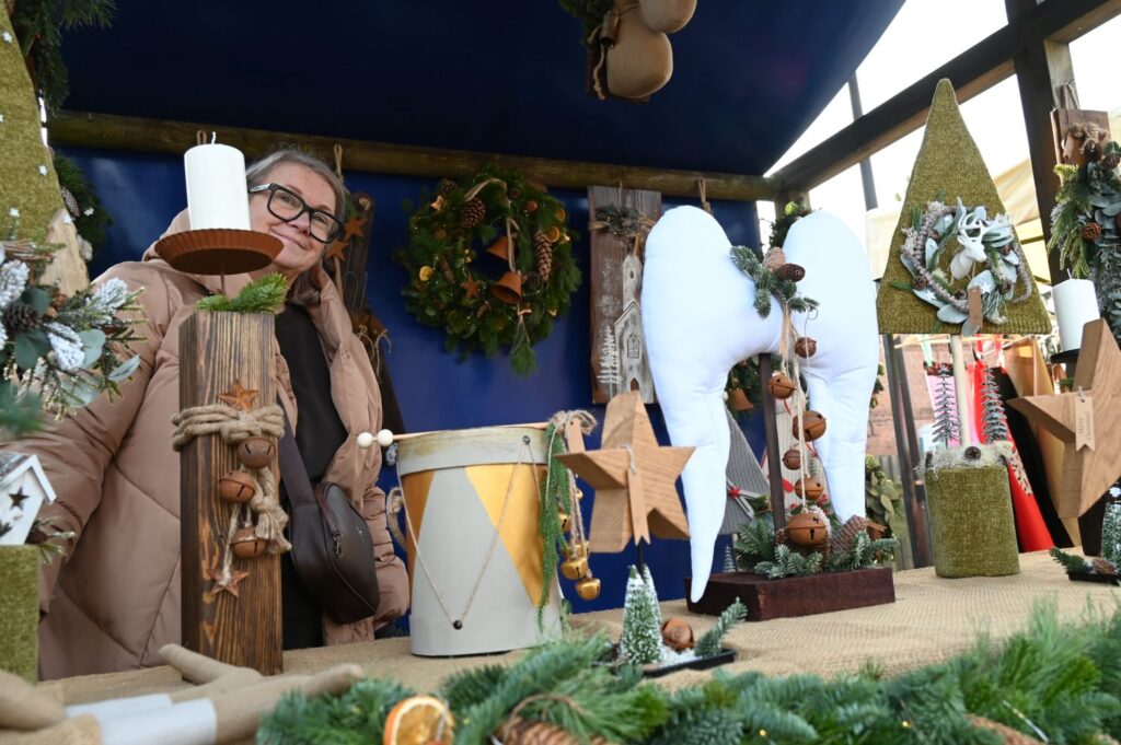 Stoisko jarmarczne z rękodziełem bożonarodzeniowym – drewniane anioły, gwiazdy i dekoracje świąteczne na tle zielonych gałązek (kliknięcie powiększy zdjęcie).