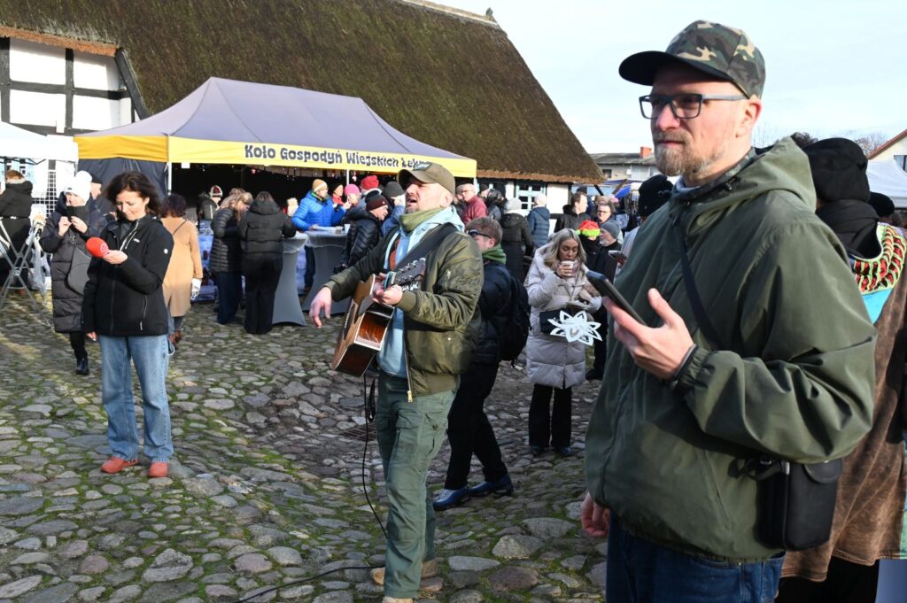 Muzyk grający na gitarze wśród uczestników jarmarku jamneńskiego na terenie skansenu, na pierwszym planie mężczyzna trzymający w ręce telefon (kliknięcie powiększy zdjęcie).